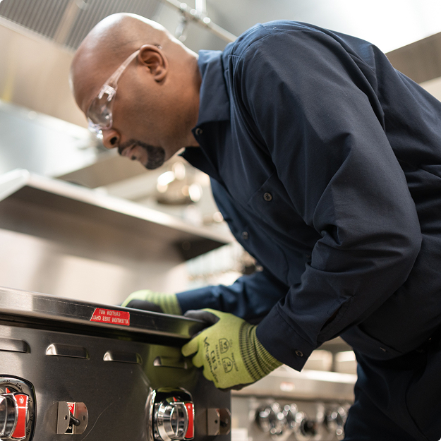Service technician in a cap and work gloves inspecting commercial kitchen equipment up close