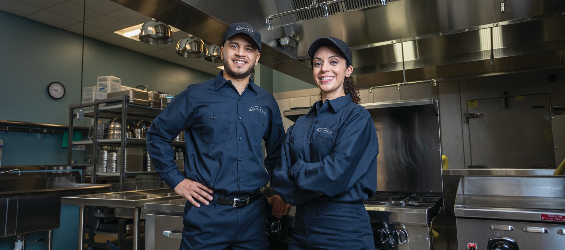 Two service technicians in navy uniforms posing with hands on hips in a commercial kitchen