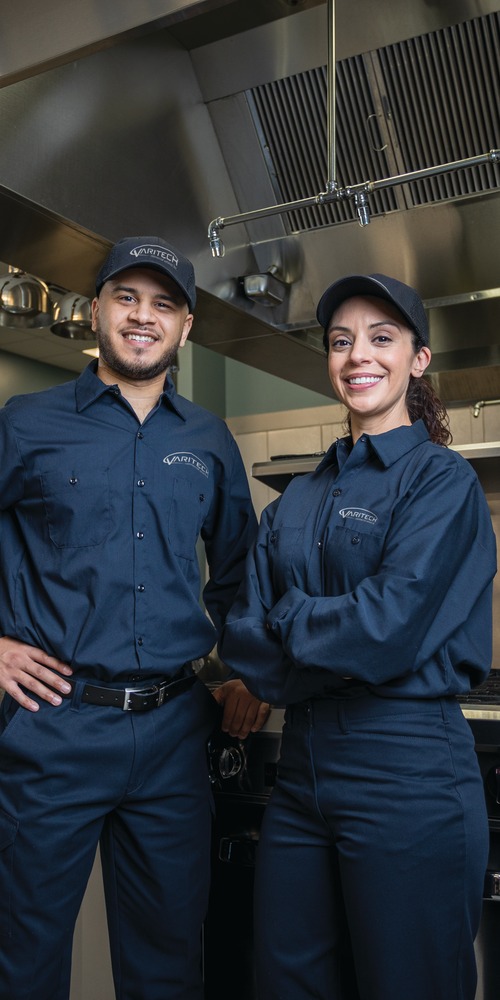 Two service technicians in navy uniforms posing with hands on hips in a commercial kitchen