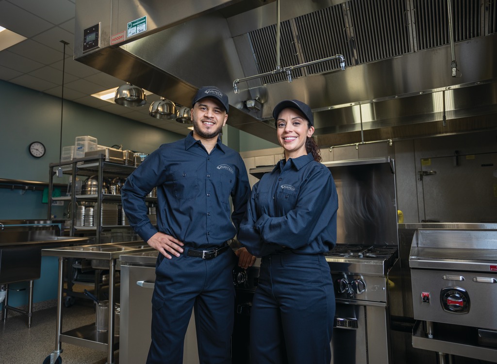 Two service technicians in navy uniforms posing with hands on hips in a commercial kitchen