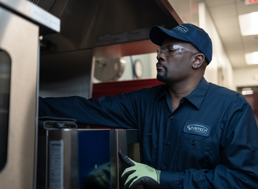 Service technician in a cap and safety glasses reaching into commercial kitchen equipment
