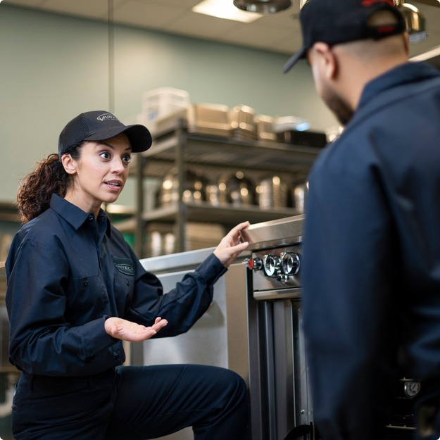 Service technician kneeling beside commercial kitchen equipment and talking with a colleague