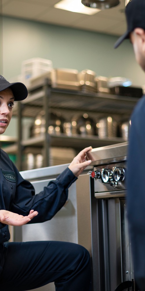 Service technician kneeling beside commercial kitchen equipment and talking with a colleague