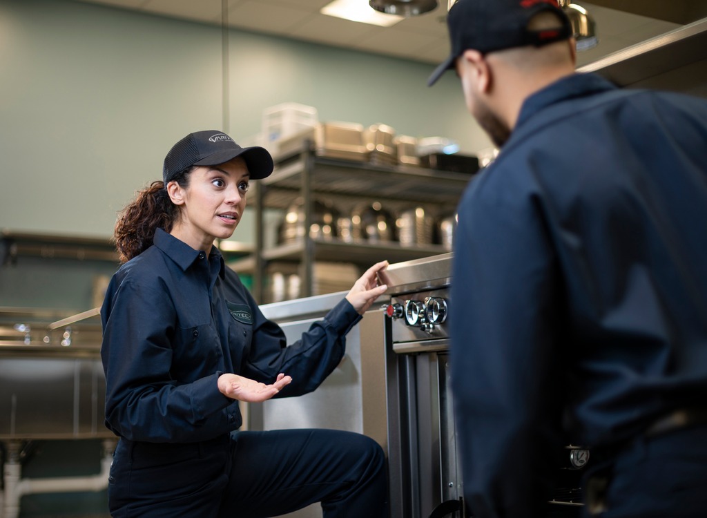 Service technician kneeling beside commercial kitchen equipment and talking with a colleague