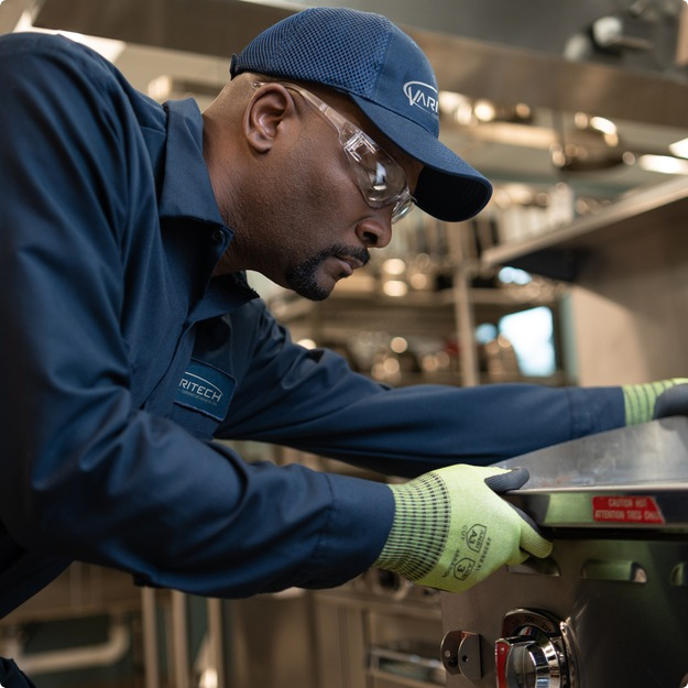 Service technician in a cap and work gloves inspecting commercial kitchen equipment up close